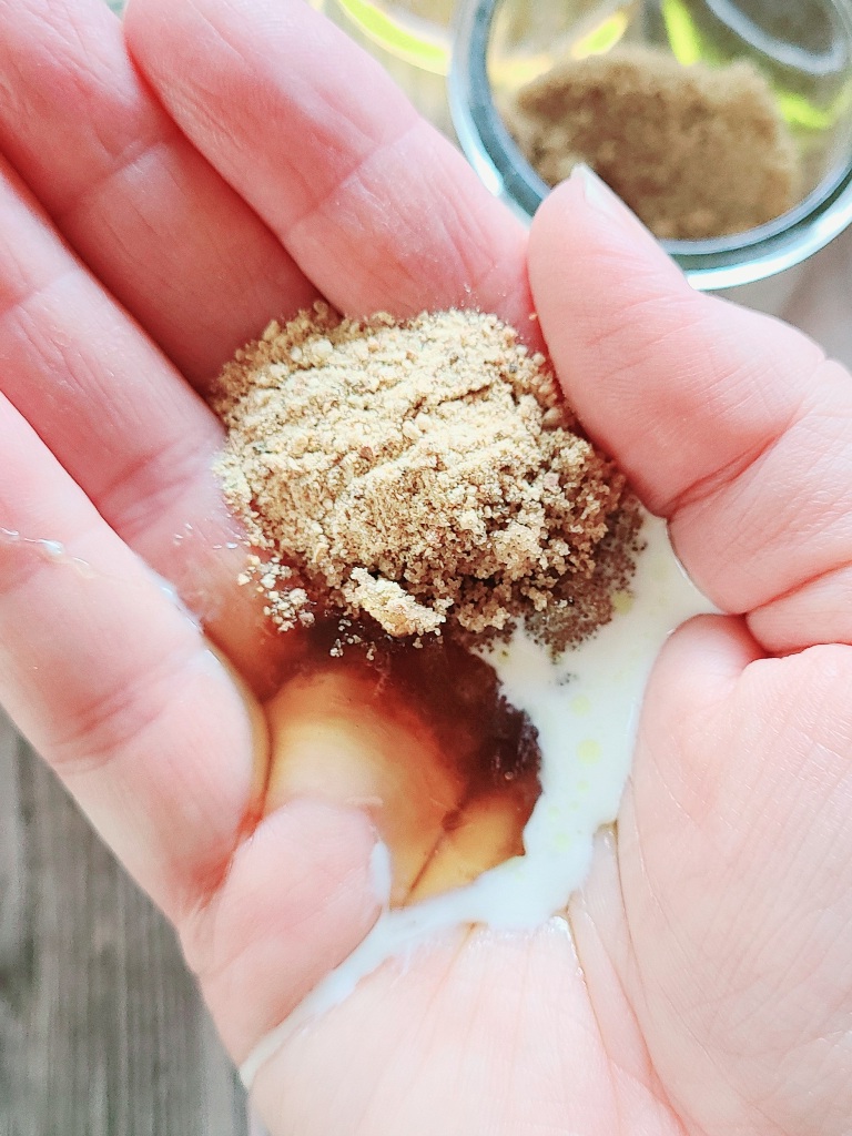Is a hand holding some brown sugar, cream, and honey in the palm and a bowl of brown sugar in the background.